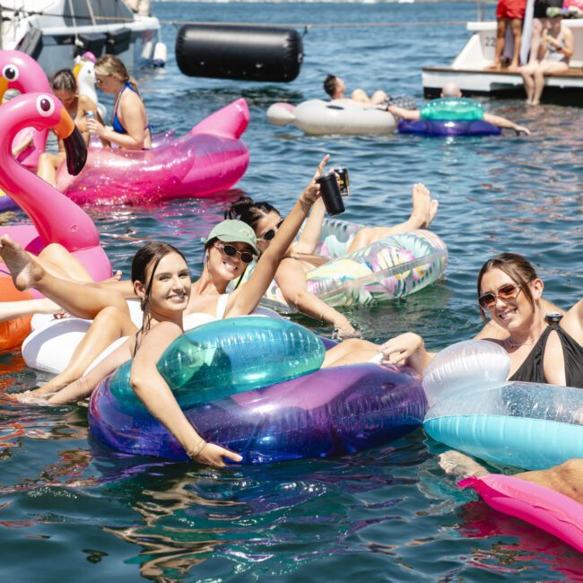 A group of people enjoying a sunny day on a lake, sitting on colorful inflatable pool floats, including a flamingo and round tubes. They are smiling, holding drinks, and surrounded by more floaters with boats in the background.