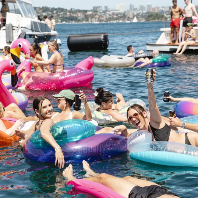 People relaxing on colorful inflatables in the water, holding drinks and smiling. Boats are anchored nearby, and a cityscape is visible in the background. The atmosphere is lively and festive.