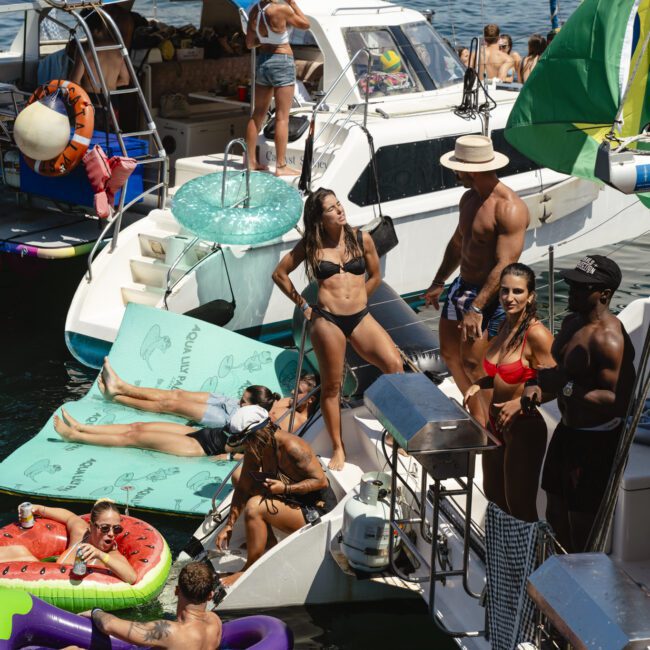 A lively scene of people enjoying a boat party on the water. They are sunbathing, lounging on inflatables, and socializing on and between several boats. Bright swimwear and sunny weather highlight the festive atmosphere.