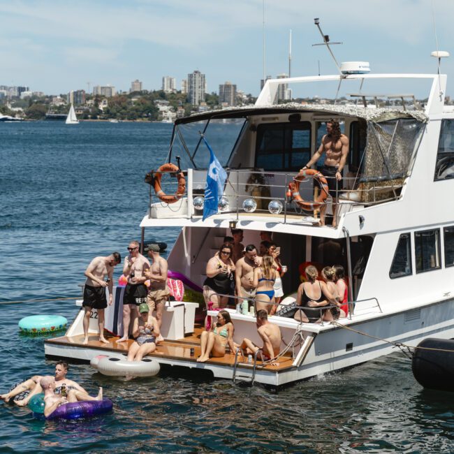 A group of people are enjoying a sunny day on a large white yacht, some are lounging on floats in the water, others are gathered on the deck. The background shows a calm blue bay with distant buildings and a few sailboats.