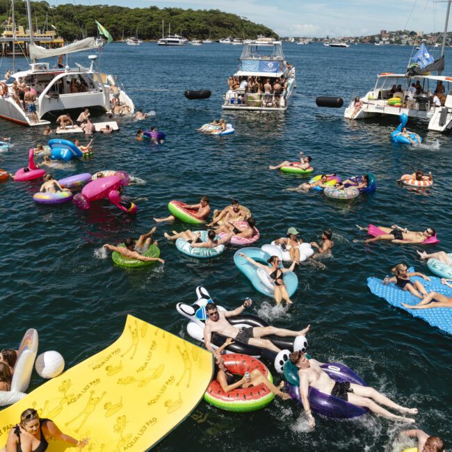 People are enjoying a sunny day on colorful inflatable floats in a bay. Several boats are anchored nearby, with more people on board. The scene is lively, with clear blue water and a forested shoreline in the background.