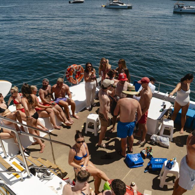 A group of people in swimwear gather on a boat deck, socializing and sunbathing under a clear blue sky. The boat is on a calm body of water with other boats visible in the background.
