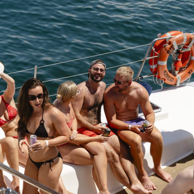 A group of people sit on a boat deck, enjoying drinks in swimsuits under a sunny sky. The ocean surrounds them, and a lifebuoy is visible on the boat's railing. They seem relaxed and smiling, with one person holding a can and another wearing sunglasses.