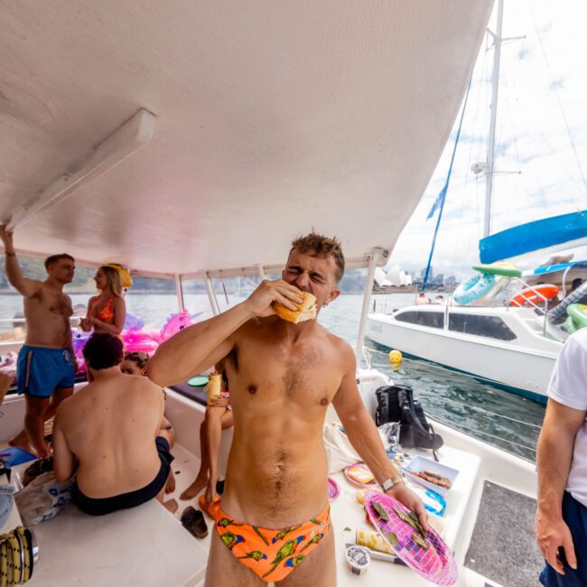 A man in patterned swimwear stands on a boat, enjoying a beverage. Other people are seated nearby, some wearing swimsuits. Another boat is visible in the background on a body of water. The scene suggests a casual, warm-day boat gathering.