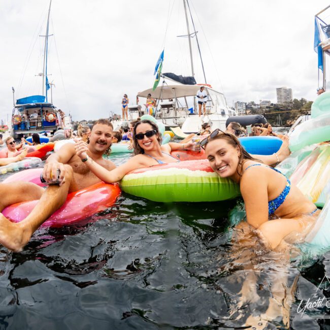 A group of people enjoying a sunny day in the water near boats. They are relaxing on colorful inflatable pool floats. The scene is lively, with many people in the background and a cityscape visible in the distance.