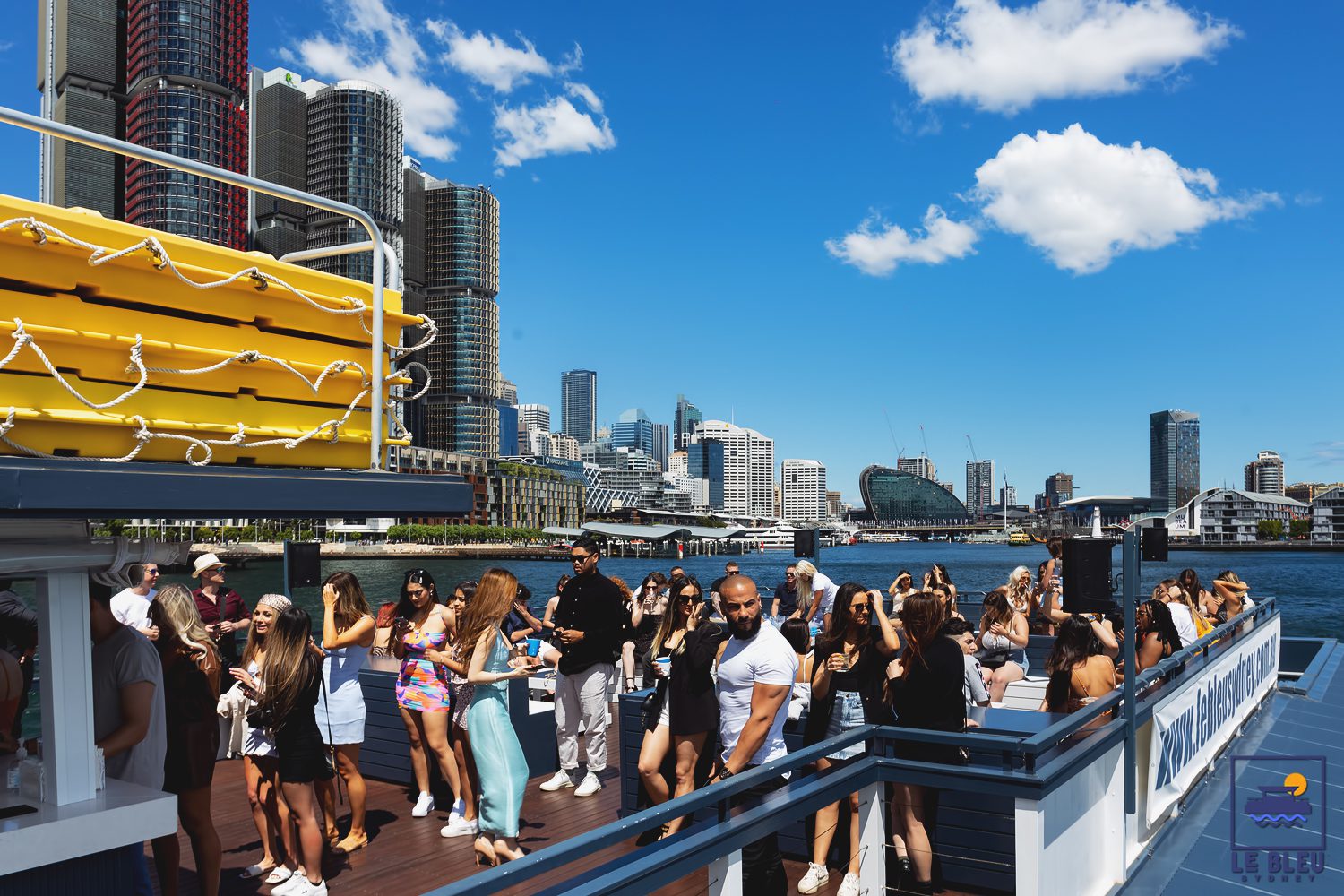 A group of people enjoys a sunny day on a boat deck with city skyscrapers and blue sky in the background, near a waterfront. Some guests are taking photos while others are mingling and relaxing.