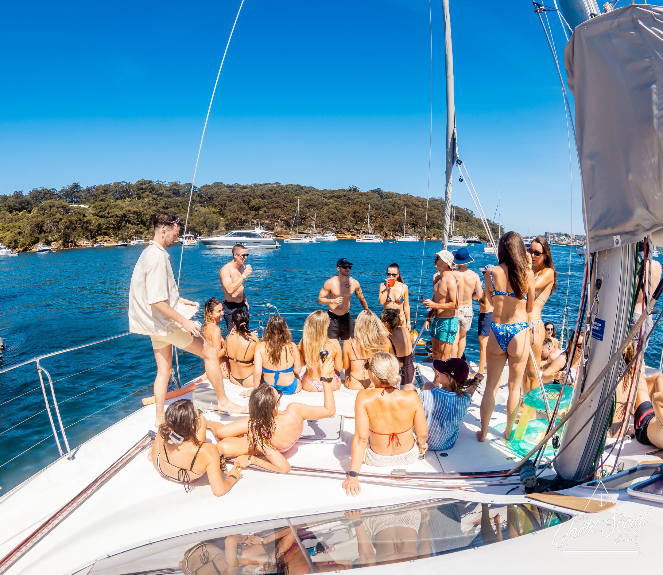A group of people in swimsuits relax and socialize on the deck of a yacht anchored in clear blue water near a wooded shoreline on a sunny day. Other boats are visible in the background.