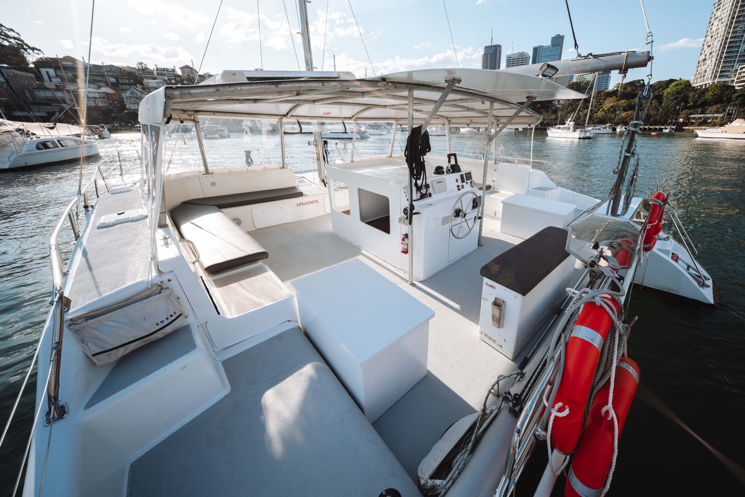 Spacious deck of a modern catamaran with seating areas, a covered helm, life preservers, and city buildings visible across the water in the background.