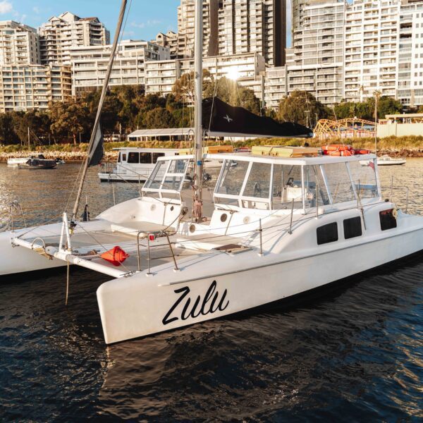 A white catamaran named "Zulu" floats on calm water near a city shoreline, with high-rise buildings and several other boats in the background on a sunny day.