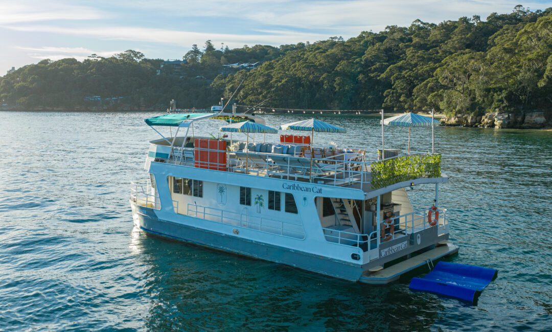 The Caribbean Cat, a gorgeous two-story white and blue vessel, drifts peacefully on tranquil water. Adorned with vibrant greenery and striped umbrellas, Caribbean Cat provides a delightful escape set before a wooded shoreline.