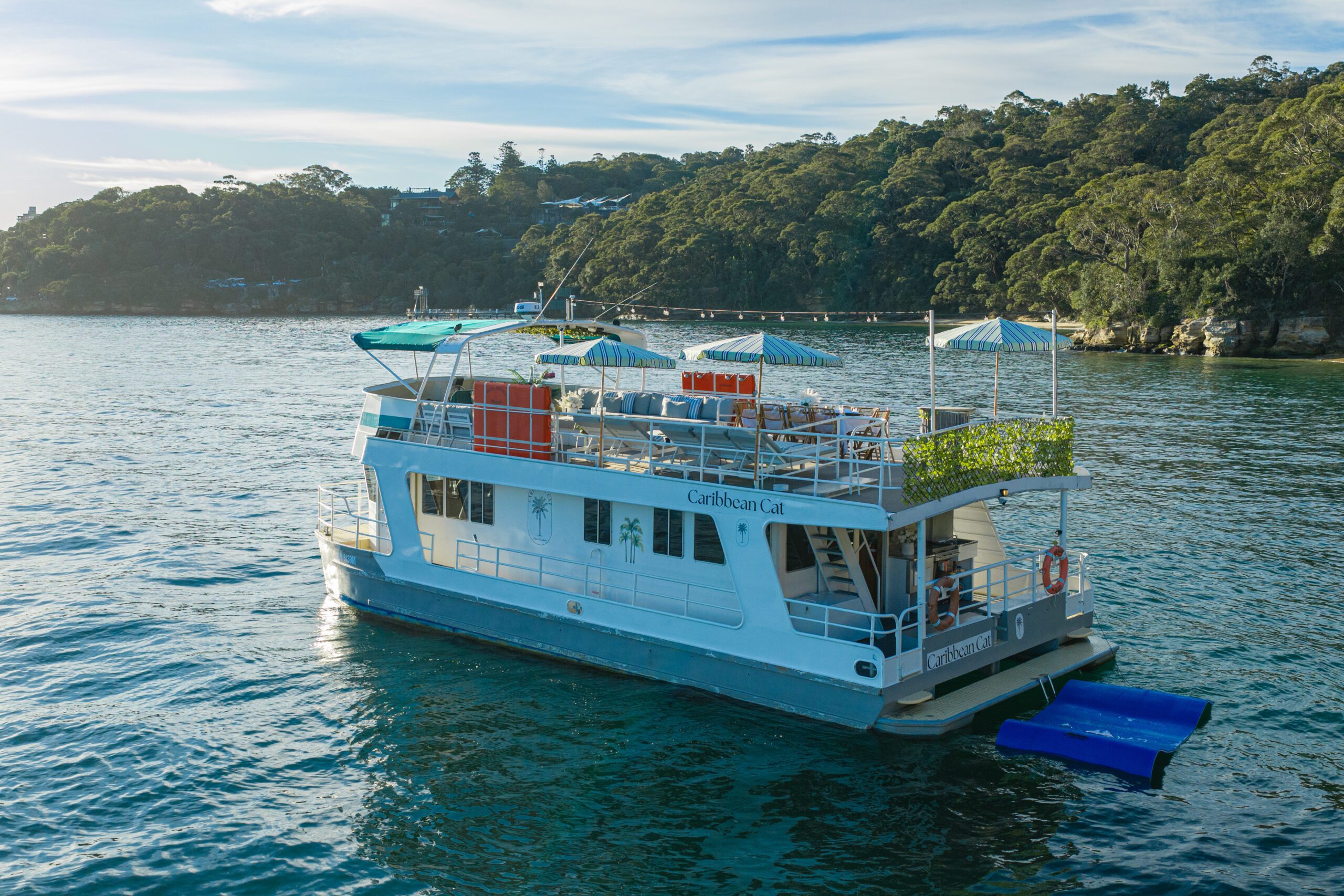 The Caribbean Cat, a gorgeous two-story white and blue vessel, drifts peacefully on tranquil water. Adorned with vibrant greenery and striped umbrellas, Caribbean Cat provides a delightful escape set before a wooded shoreline.