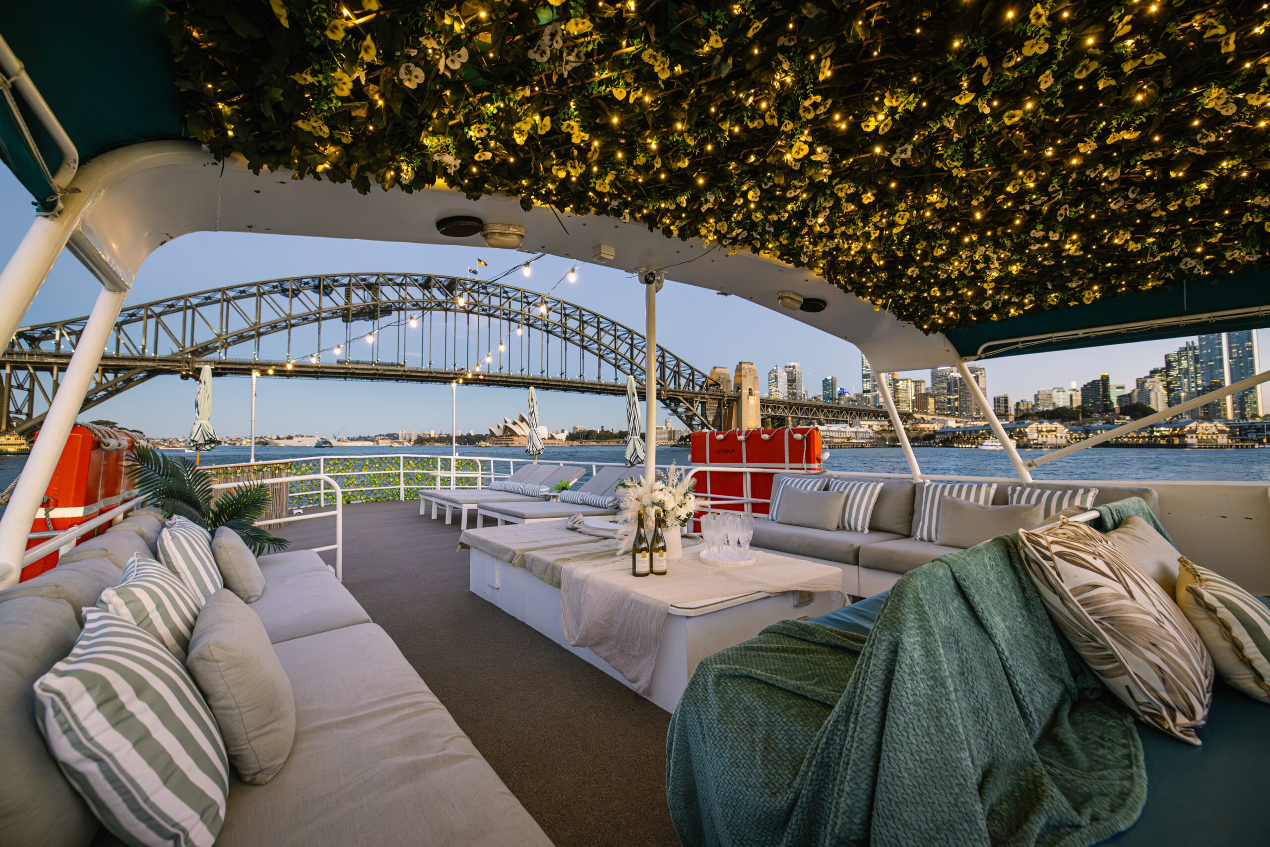 Experience luxury on the Caribbean Cat’s boat deck, featuring cozy sofas, striped pillows, floral ceiling lights, and a table set for drinks—all while taking in breathtaking sunset views of Sydney Harbour Bridge and the city skyline.