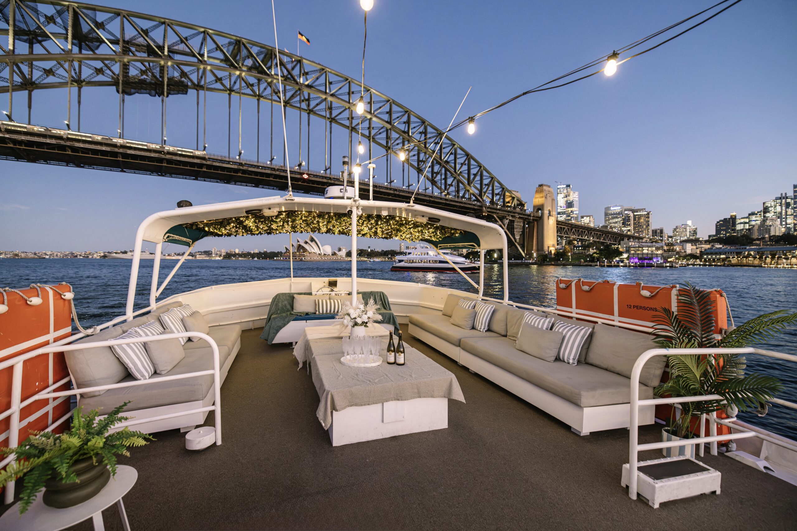 The Caribbean Cat—a cozy boat lounge featuring sofas and string lights—floats on the water near Sydney Harbour Bridge at sunset, with city buildings and the Opera House visible in the background.
