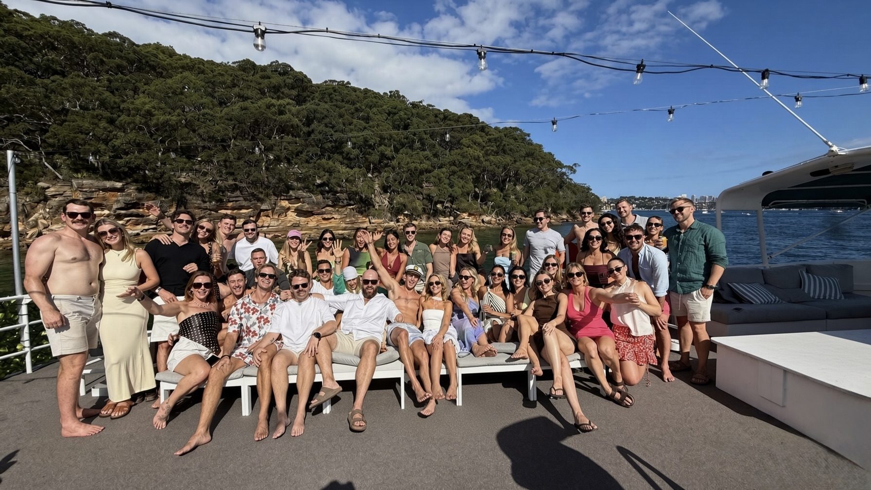 A large group of people pose and smile on a boat deck with the Caribbean Cat, surrounded by trees, a rocky shoreline, and a blue sky with clouds in the background.