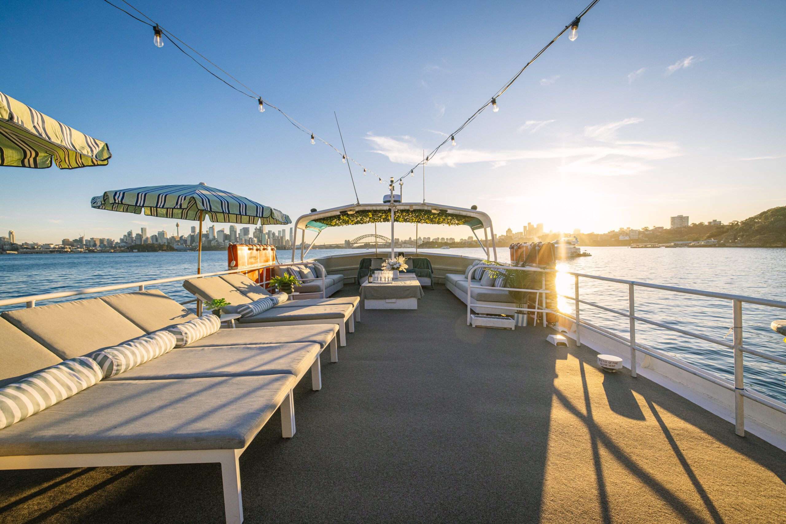 Loungers and tables on a boat deck under striped umbrellas, with string lights overhead, overlook calm water and a city skyline at sunset—perfectly capturing the laid-back charm of the Caribbean Cat experience.