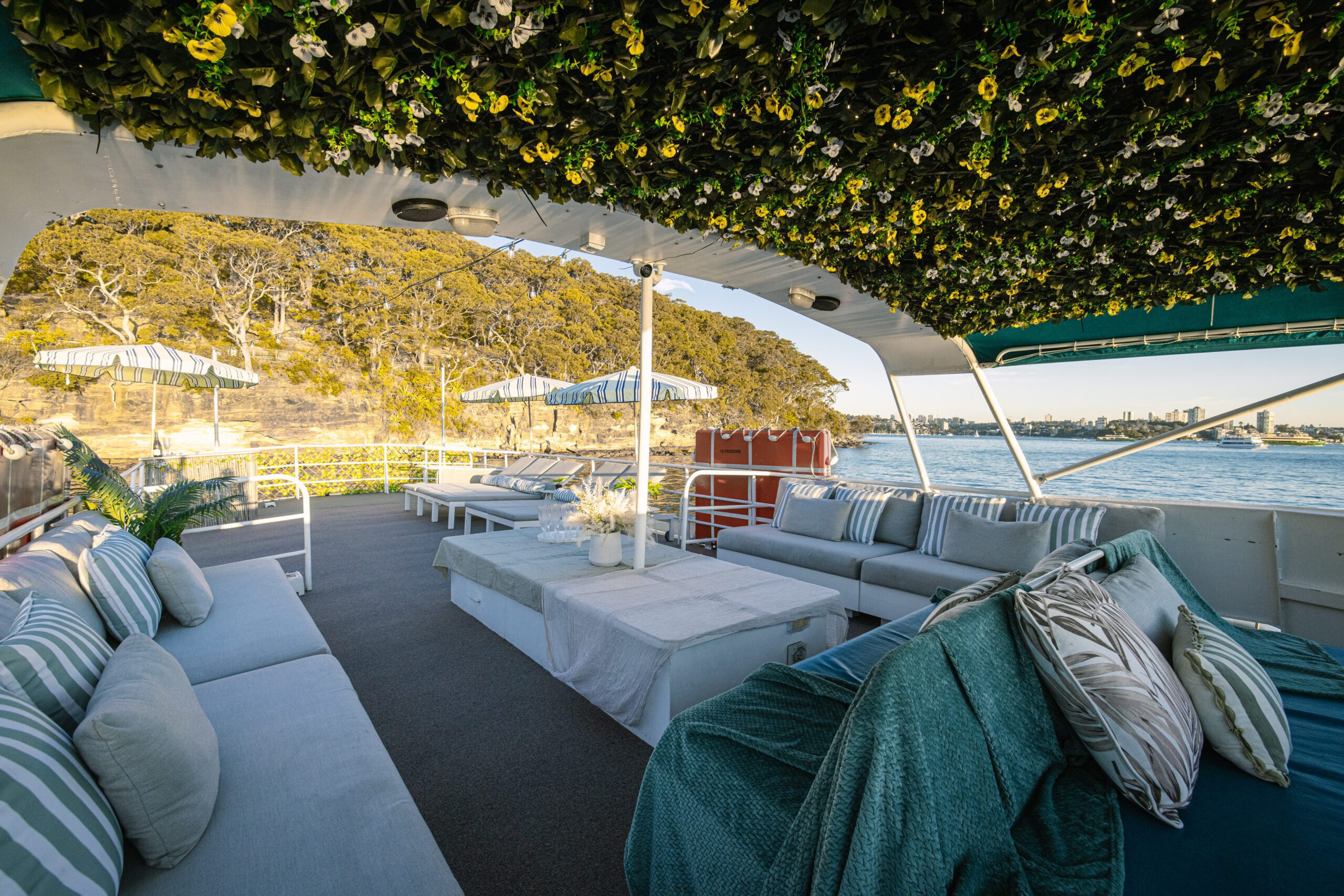 Experience the Caribbean Cat: a boat deck featuring cushioned seating, striped pillows, parasols, and a ceiling draped in lush green foliage with yellow flowers—an idyllic oasis for relaxing and unwinding. Enjoy views of tranquil water and trees in the background for the ultimate getaway.