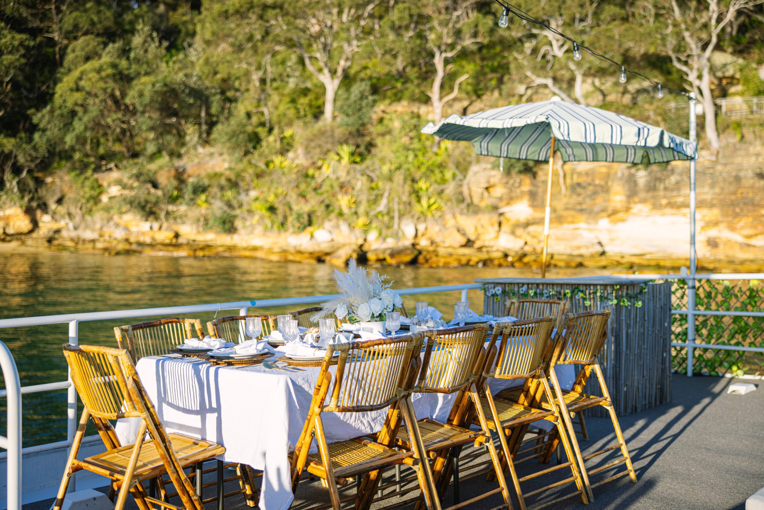 A table with a white cloth and bamboo chairs is set for a meal on a deck by the water, as the Caribbean Cat looks on curiously beneath the shade of a striped umbrella, while trees sway gently in the background.