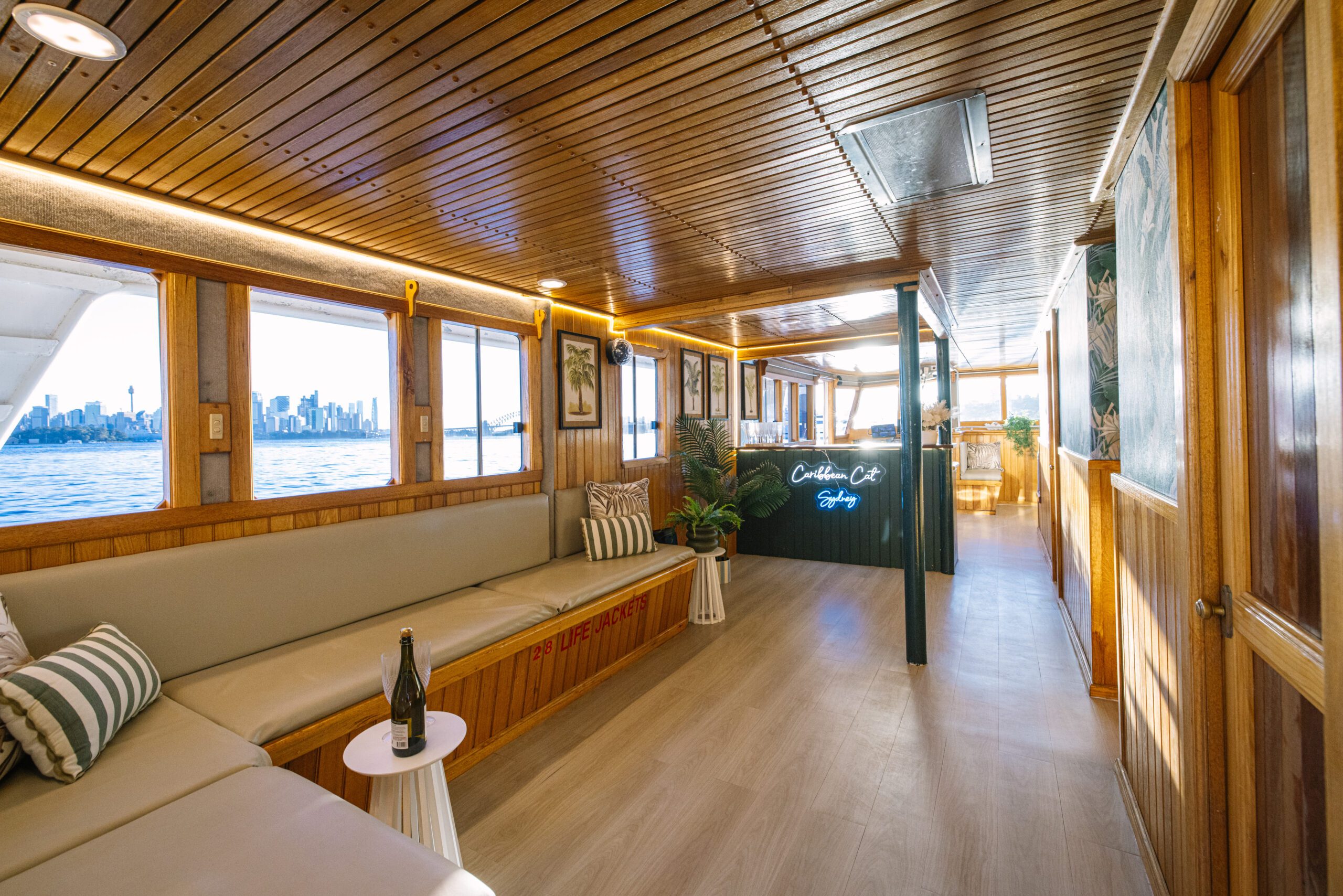 A boat’s wood-paneled interior showcases beige seating, striped pillows, a small table with a bottle, decorative plants, expansive windows overlooking a city skyline, and a sign that reads “Caribbean Cat,” creating a cozy atmosphere inspired by the relaxed charm of the Caribbean Cat product.