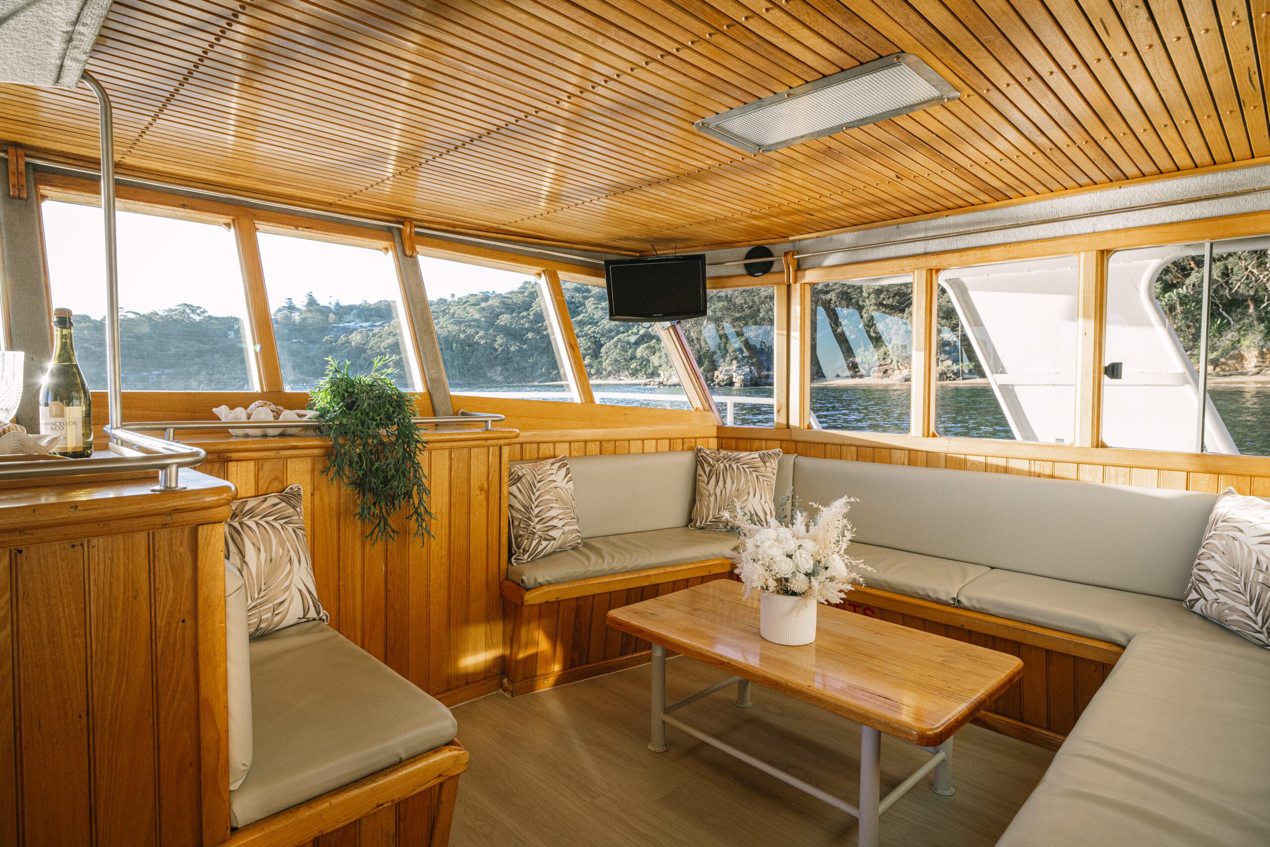 A cozy boat lounge with wooden walls and ceiling, beige cushioned benches, a wooden coffee table adorned with a white flower arrangement, expansive windows, and scenic outdoor views—a tranquil retreat embodying the signature comfort and inviting atmosphere of the Caribbean Cat.
