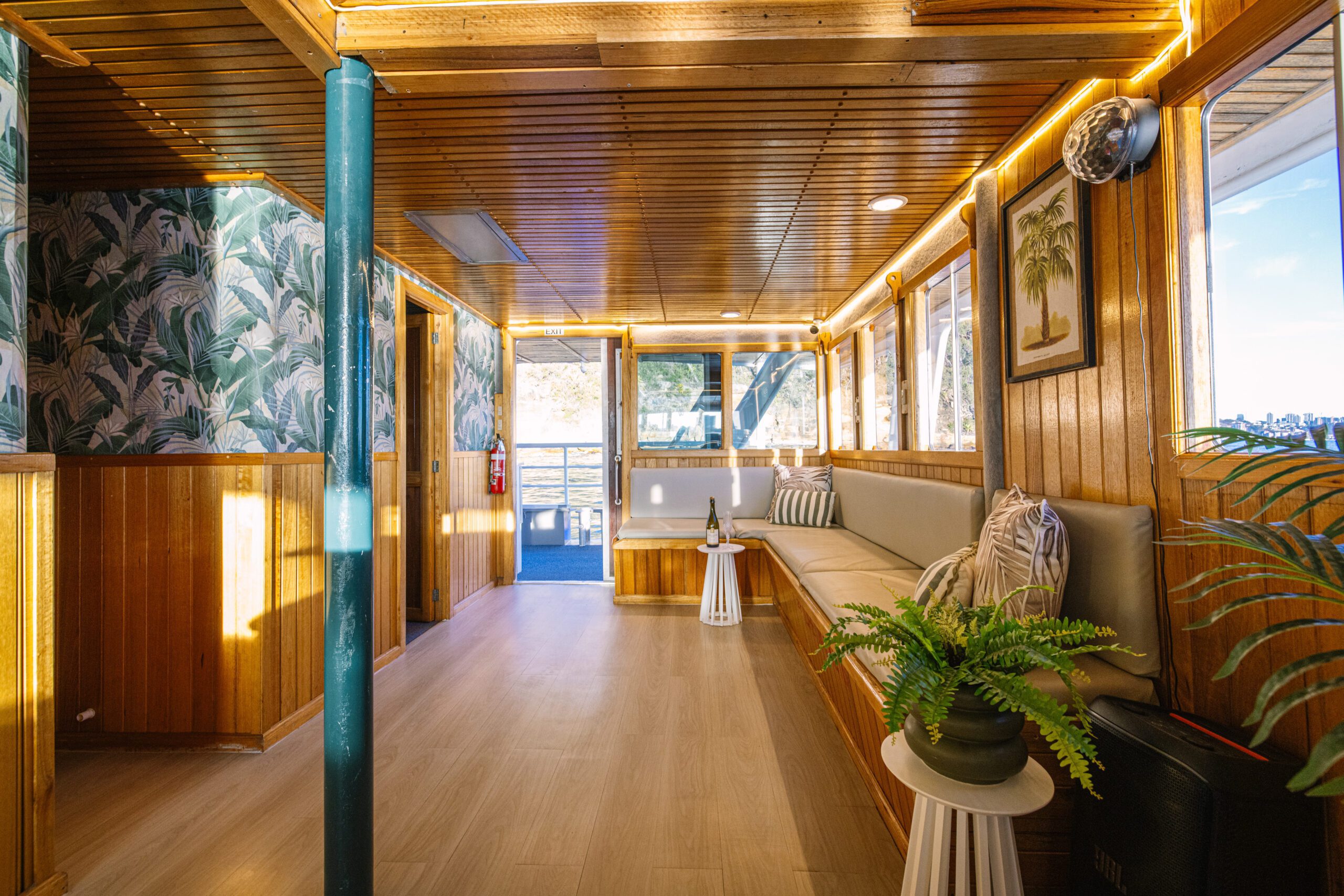 Step inside the Caribbean Cat to enjoy a cozy interior featuring elegant wood paneling, vibrant tropical wallpaper, plush cushioned seating, potted plants for a touch of greenery, convenient small tables, and expansive windows that bathe the space in sunlight while offering stunning views of the water outside.