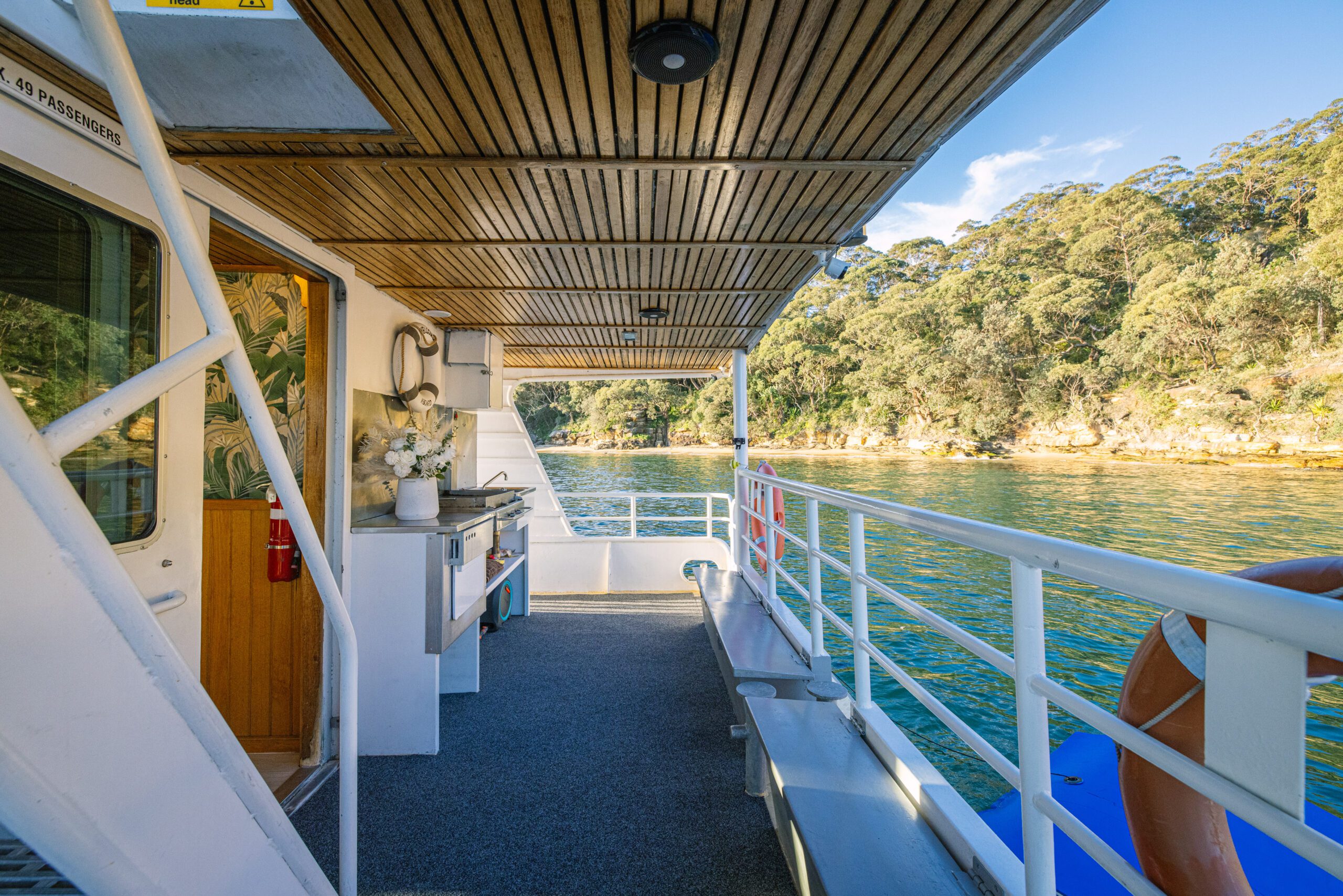 A Caribbean Cat boat deck, featuring comfortable benches, a lifebuoy, and a compact kitchenette, overlooks serene sunlit green waters and a lush tree-lined shore beneath a partly cloudy sky—an inviting scene evocative of tranquil cruises on the Caribbean Cat.