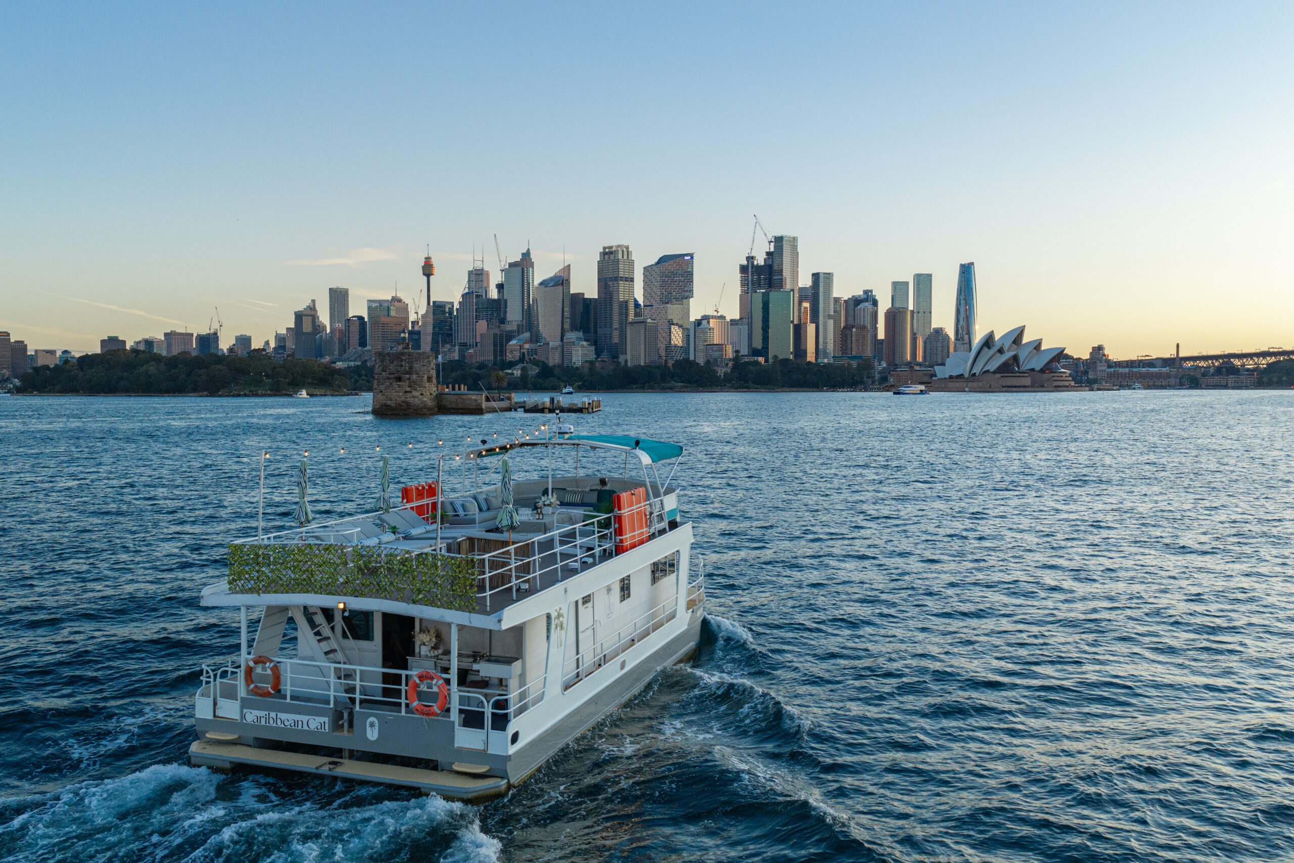 A Caribbean Cat ferry glides across Sydney Harbour at sunset, with the Sydney Opera House, the city skyline, and the Harbour Bridge visible in the background.