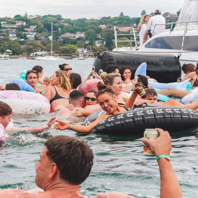 A group of people in swimsuits float on colorful inflatables and swim in the water near a yacht. They are smiling, laughing, and enjoying drinks, with houses and greenery visible in the background.