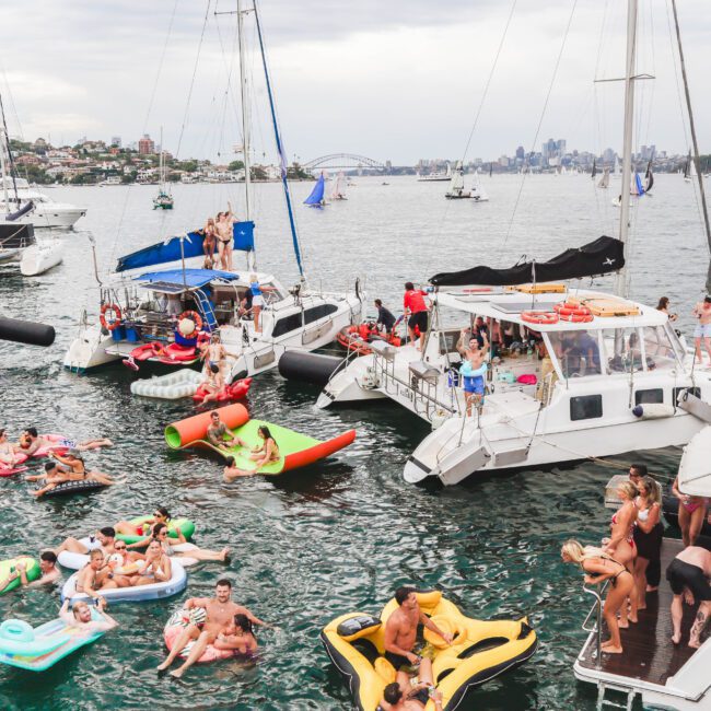 People gather on boats and colorful inflatables in a busy harbor, enjoying a lively party on the water. The background shows a city skyline and the Sydney Harbour Bridge under a cloudy sky.