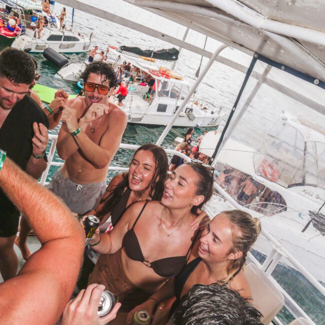 A group of young adults in swimsuits smile and dance on a boat, holding drinks. Other boats and people are visible on the water in the background, suggesting a lively party atmosphere.