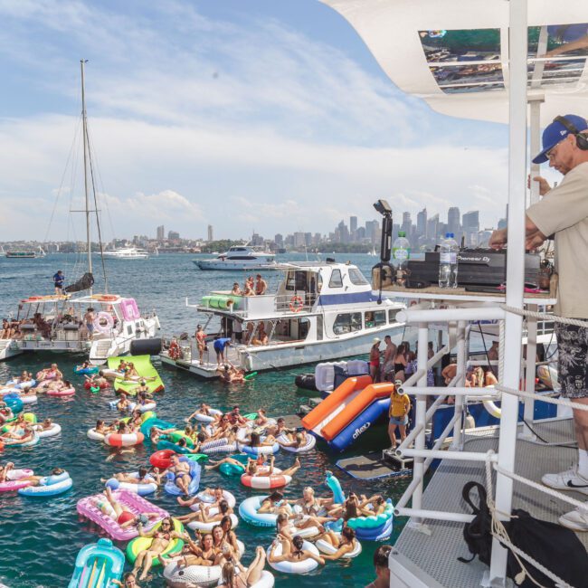 A DJ plays music on a boat while people relax on colorful inflatable floats in the water and gather on boats nearby, with a city skyline visible in the background on a sunny day.