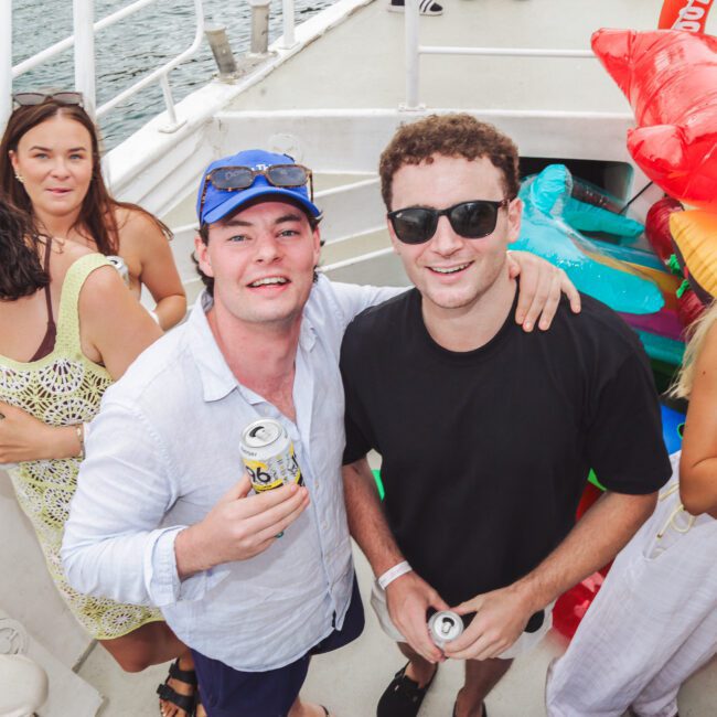 A group of young adults enjoy a boat party, holding drinks and smiling. Inflatable pool floats are stacked in the background, and the boat is surrounded by water. The mood is lively and festive.