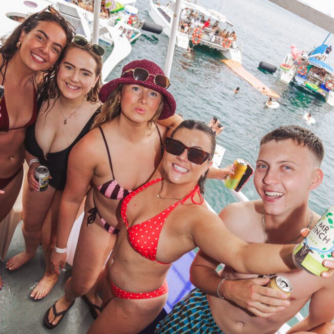 A group of five young adults in swimsuits smile and pose with canned drinks on a boat, with other boats and people swimming in the ocean in the background. The mood is festive and summery.