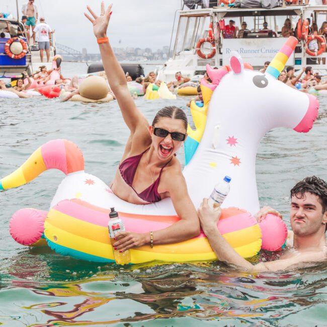 A woman in sunglasses and a maroon bikini sits on a colorful inflatable unicorn in the water, smiling and raising her arm, while a man next to her holds the float. Boats and people are visible in the background.