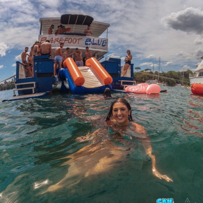 A woman smiles while swimming in clear water near a party boat with a slide. Several people are on the boat, and the boat’s name, "Barefoot Blue," is visible. The sky is partly cloudy. Logos say "Sail Gustavo" and "Lighthouse Social.