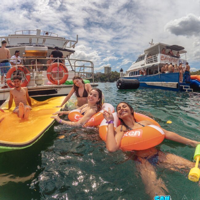 Four people float on orange and white inflatable rings in the water near two boats under a partly cloudy sky. Two women smile at the camera while others relax nearby. The scene is lively and summery.