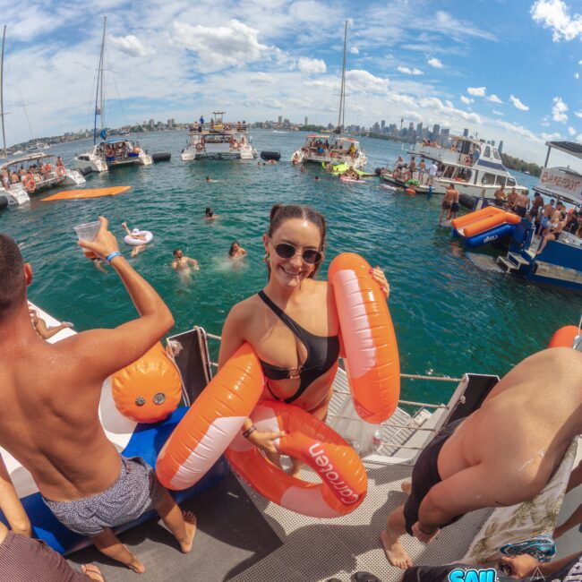 A woman in sunglasses and a bikini smiles at the camera while holding inflatable pool floats on a boat. People swim and relax on boats in a lively scene on the water with a city skyline in the background.