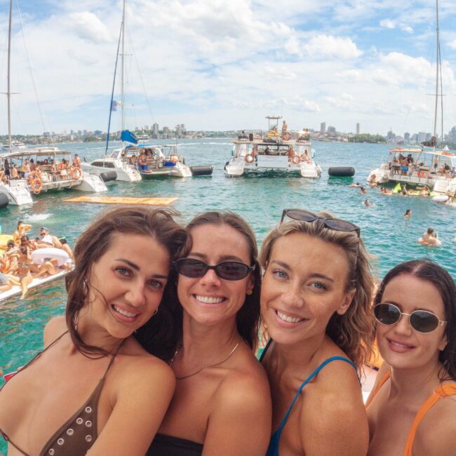 Four women in swimsuits smile and pose on a boat surrounded by water, with several other boats and people in the background. The sky is partly cloudy, and a city skyline is visible in the distance.