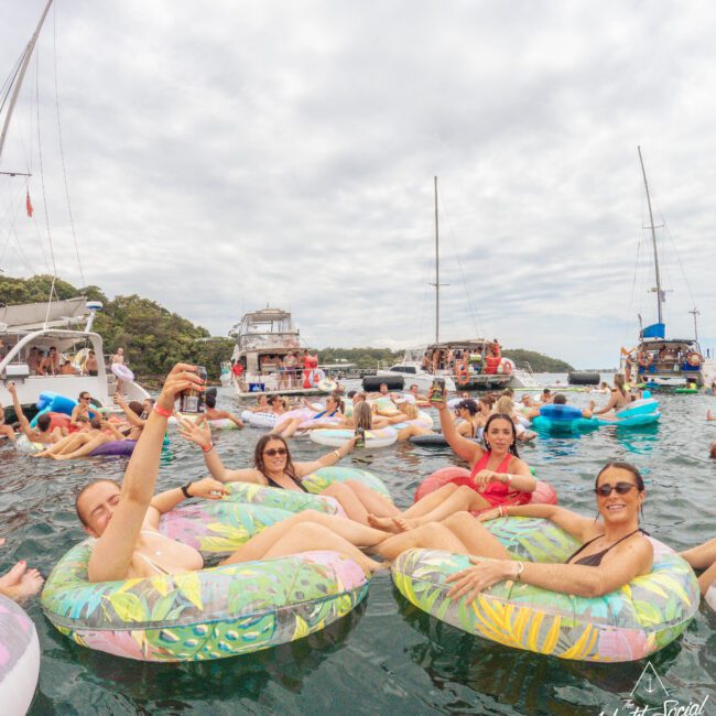 A group of people float on colorful pool inflatables in the water, smiling and holding drinks, with several boats and others enjoying the scene in the background under a cloudy sky.
