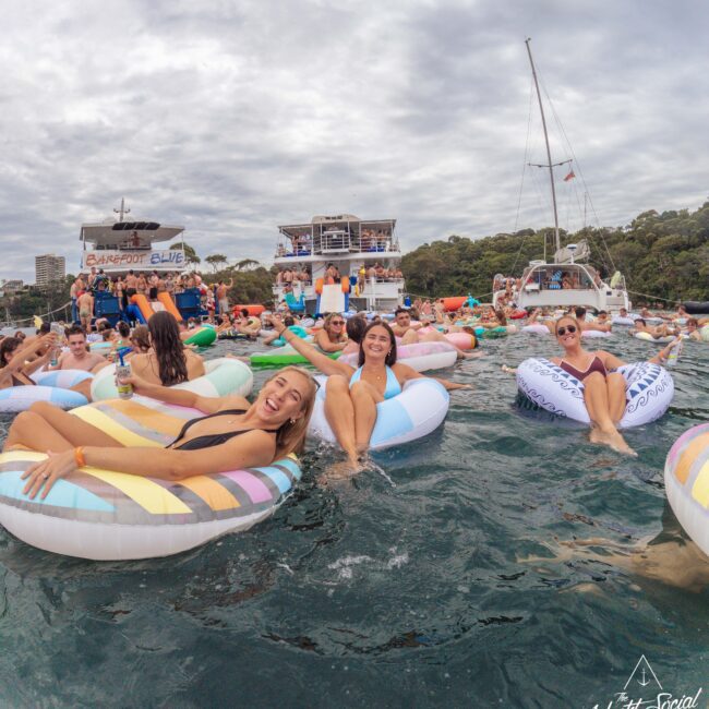 A group of people relax on colorful inflatable pool floats in open water near boats, smiling and enjoying a social gathering under a cloudy sky with green trees in the background.
