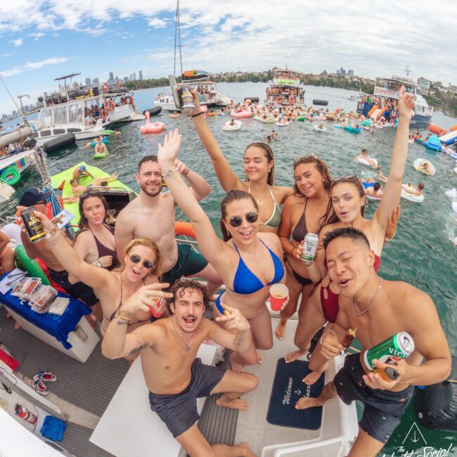 A group of young adults in swimsuits smile, cheer, and hold drinks on a boat during a lively summer party. Other boats and people floating in the water are visible in the background under a partly cloudy sky.