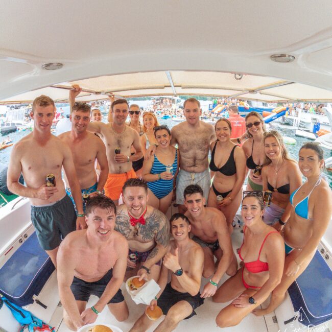 A group of people in swimsuits smile and pose together on a boat, some holding drinks or food. The boat is crowded and other boats with people are visible in the background on the water.