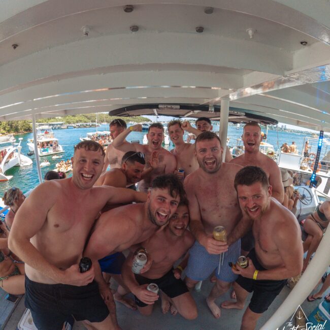 A group of shirtless men smile and pose with drinks on a crowded party boat, with other boats and people visible in the water behind them on a sunny day.
