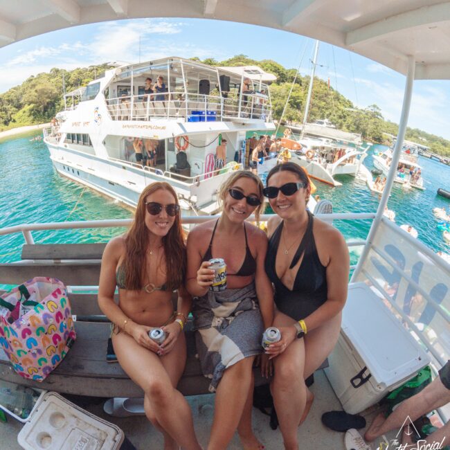 Three women in swimsuits sit on a boat bench, smiling and holding drinks. Behind them are other boats on clear blue water with people enjoying the sunny day. A cooler and colorful bag are beside them. Trees are visible in the background.
