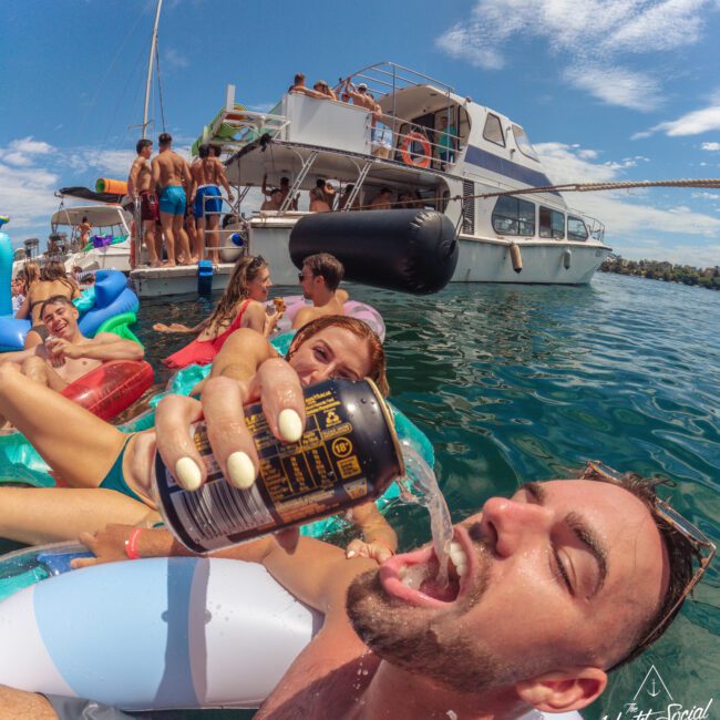 A man on a float pours a drink into his mouth from a can while lounging with friends on colorful inflatables in the water; a party boat with more people is seen in the background.
