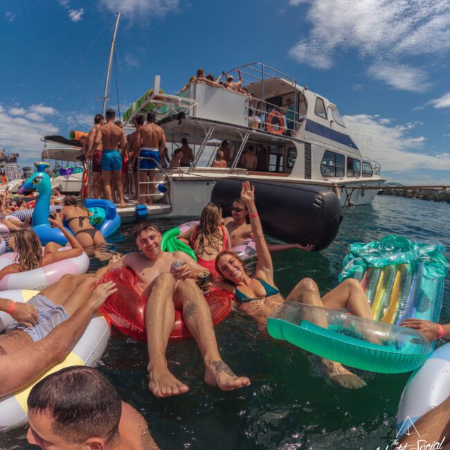 People relax on inflatable floats in the water near a large yacht, enjoying a sunny day. Some wave at the camera, while others chat or lounge. The scene is lively with bright skies and festive atmosphere.