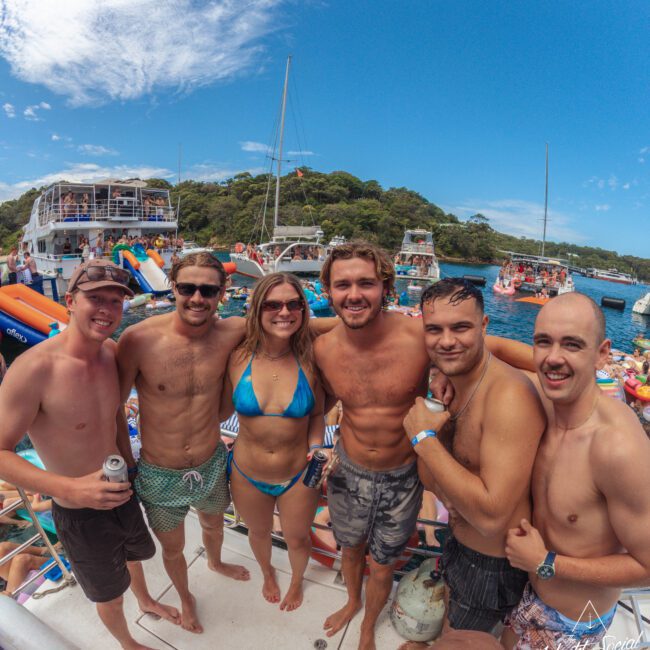 A group of six smiling friends, four men and two women in swimsuits, pose together on a crowded boat under a sunny sky, with other boats, people, and lush green hills in the background.