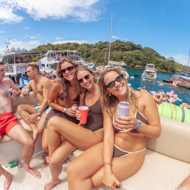 Three women in swimsuits smile and pose with drinks on a boat, surrounded by other people and boats on clear blue water under a sunny sky; a festive, summery atmosphere.