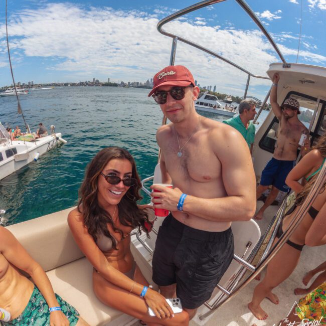 A group of young adults enjoy a sunny day on a boat. A smiling woman in sunglasses sits next to a man in a red cap holding drinks. Other people relax and socialize in swimwear, with water and boats in the background.