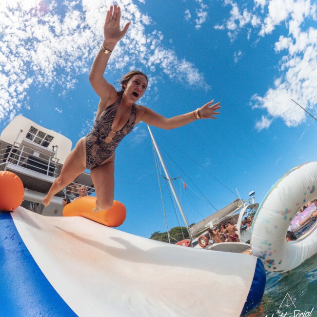 A woman in a patterned swimsuit slides down an inflatable water slide from a boat, arms up and mouth open, with people watching and blue sky with clouds in the background. The scene is energetic and playful.