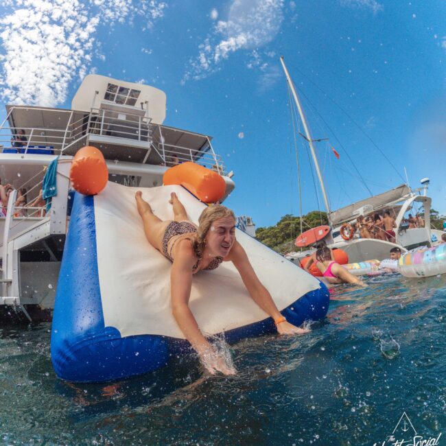 A woman in a swimsuit slides down an inflatable water slide from a yacht into the ocean, with other boats and people in the background under a blue sky with scattered clouds.